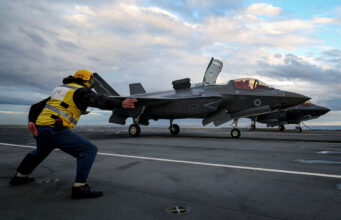 Crew member in a yellow safety vest pushes a grey F-35 fighter on a crowded aircraft carrier deck under a cloudy sky, cockpit canopy open nearby.