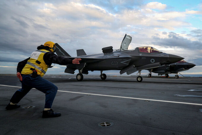British aircraft carrier launches fighter jets in Arctic Crew member in a yellow safety vest pushes a grey F-35 fighter on a crowded aircraft carrier deck under a cloudy sky, cockpit canopy open nearby.