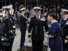 Royal Navy commissions minehunting mothership Naval officers in dress uniforms stand in formation as a woman in a dark dress speaks with them during a formal ceremony.