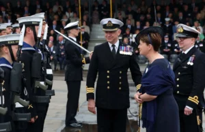 Royal Navy commissions minehunting mothership Naval officers in dress uniforms stand in formation as a woman in a dark dress speaks with them during a formal ceremony.
