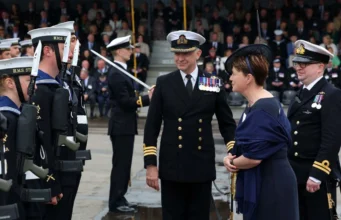 Royal Navy commissions minehunting mothership Naval officers in dress uniforms stand in formation as a woman in a dark dress speaks with them during a formal ceremony.