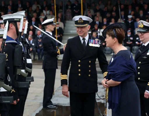 Naval officers in dress uniforms stand in formation as a woman in a dark dress speaks with them during a formal ceremony.