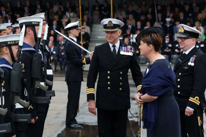 Royal Navy commissions minehunting mothership Naval officers in dress uniforms stand in formation as a woman in a dark dress speaks with them during a formal ceremony.
