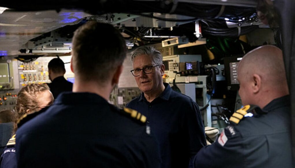 An older man with glasses speaks to a group of uniformed sailors inside a technical control room or ship compartment.