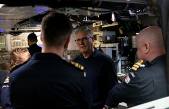 An older man with glasses speaks to a group of uniformed sailors inside a technical control room or ship compartment.