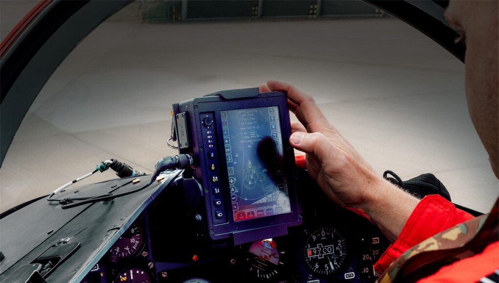 GECO x Red Arrows #4 Pilot in a red flight suit uses a rugged tablet-style display in the fighter jet cockpit, surrounded by instrument gauges.