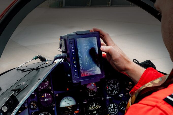 Pilot in a red flight suit uses a rugged tablet-style display in the fighter jet cockpit, surrounded by instrument gauges.