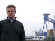 Man in a dark gray sweater stands outdoors at a shipyard with a large blue gantry crane in the background.
