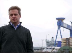 Man in a dark gray sweater stands outdoors at a shipyard with a large blue gantry crane in the background.