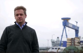 Man in a dark gray sweater stands outdoors at a shipyard with a large blue gantry crane in the background.