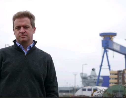 Man in a dark gray sweater stands outdoors at a shipyard with a large blue gantry crane in the background.