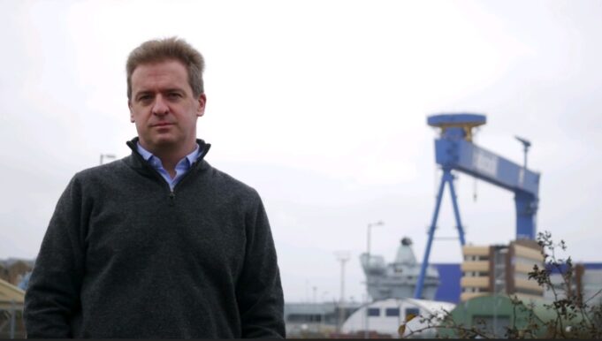 Man in a dark gray sweater stands outdoors at a shipyard with a large blue gantry crane in the background.
