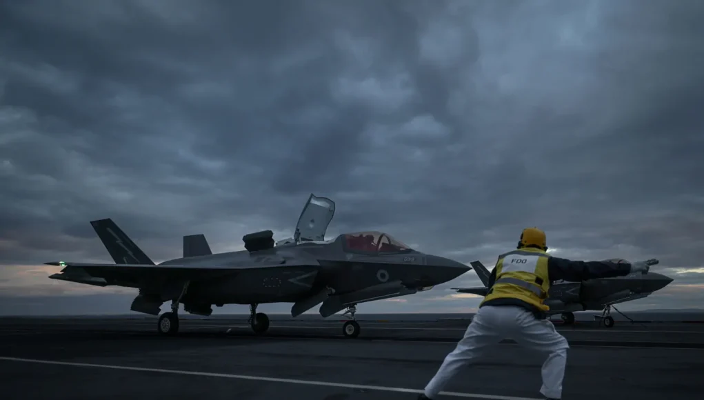 Its go for launch from the Flight Deck Officer Two fighter jets parked on a dark aircraft carrier deck at dusk, with a crew member in a yellow vest guiding them.
