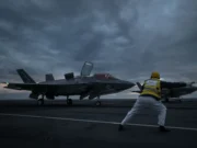 British airpower flexes muscle near Russia Two fighter jets parked on a dark aircraft carrier deck at dusk, with a crew member in a yellow vest guiding them.