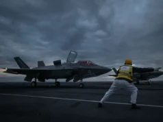 British airpower flexes muscle near Russia Two fighter jets parked on a dark aircraft carrier deck at dusk, with a crew member in a yellow vest guiding them.