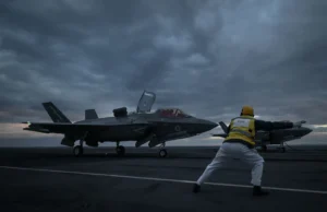 British airpower flexes muscle near Russia Two fighter jets parked on a dark aircraft carrier deck at dusk, with a crew member in a yellow vest guiding them.