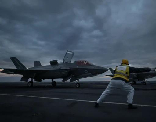 Two fighter jets parked on a dark aircraft carrier deck at dusk, with a crew member in a yellow vest guiding them.