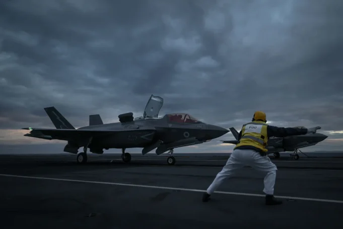 Two fighter jets parked on a dark aircraft carrier deck at dusk, with a crew member in a yellow vest guiding them.
