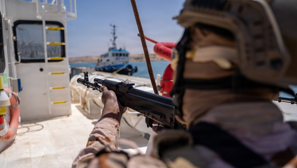 LIBYAN Soldier in camouflage aiming a rifle from the deck of a ship, with a small patrol boat visible in the water in the background.