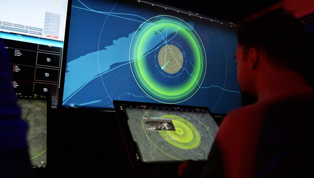 People in a dark control room monitor a large radar/map display with bright green concentric rings on a wall-sized screen