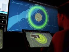 People in a dark control room monitor a large radar/map display with bright green concentric rings on a wall-sized screen