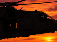 Silhouette of a military helicopter flying at sunset against a fiery orange sky.