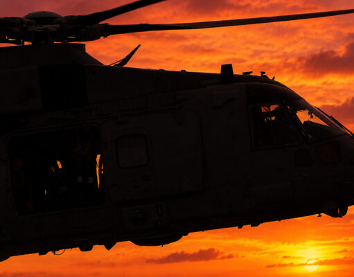 Silhouette of a military helicopter flying at sunset against a fiery orange sky.