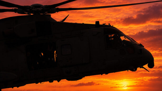 Silhouette of a military helicopter flying at sunset against a fiery orange sky.