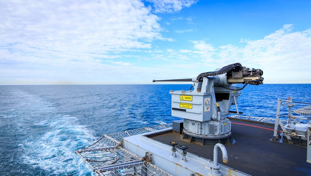 Naval gun mounted on a ship's deck with the open ocean and blue sky in the background.