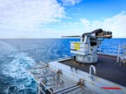Naval gun mounted on a ship's deck with the open ocean and blue sky in the background.