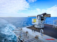 Naval gun mounted on a ship's deck with the open ocean and blue sky in the background.