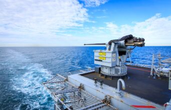 Naval gun mounted on a ship's deck with the open ocean and blue sky in the background.