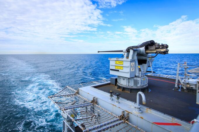 Naval gun mounted on a ship's deck with the open ocean and blue sky in the background.