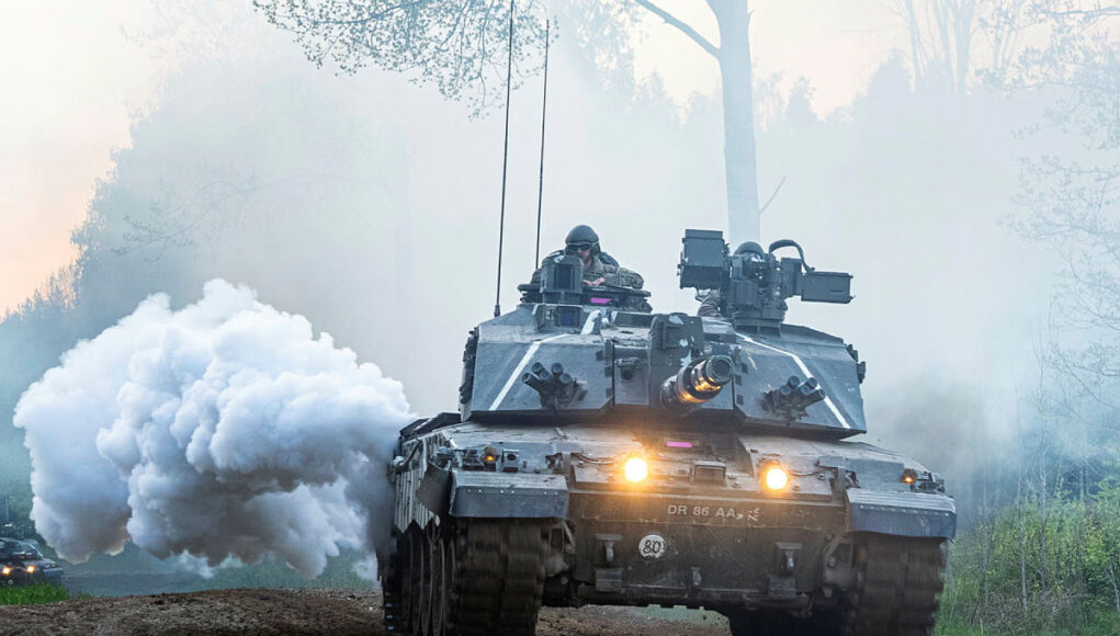 OP_CABRIT_ExSPST_20260521_FLF_Lt_Olivier.jpg.iCf_2sCBMrGTPQNlYBMA.K1NI8Fqg7H Armored battle tank driving on a dirt road through a forest, with smoke plume and headlights lit, soldiers visible in the turret.