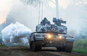 Armored battle tank driving on a dirt road through a forest, with smoke plume and headlights lit, soldiers visible in the turret.