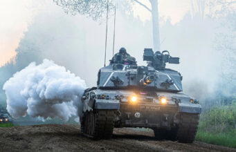 Armored battle tank driving on a dirt road through a forest, with smoke plume and headlights lit, soldiers visible in the turret.