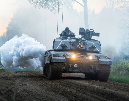 Armored battle tank driving on a dirt road through a forest, with smoke plume and headlights lit, soldiers visible in the turret.