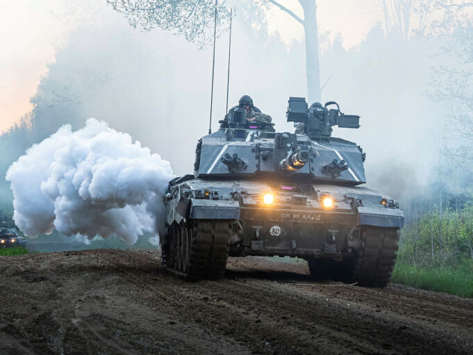 Armored battle tank driving on a dirt road through a forest, with smoke plume and headlights lit, soldiers visible in the turret.