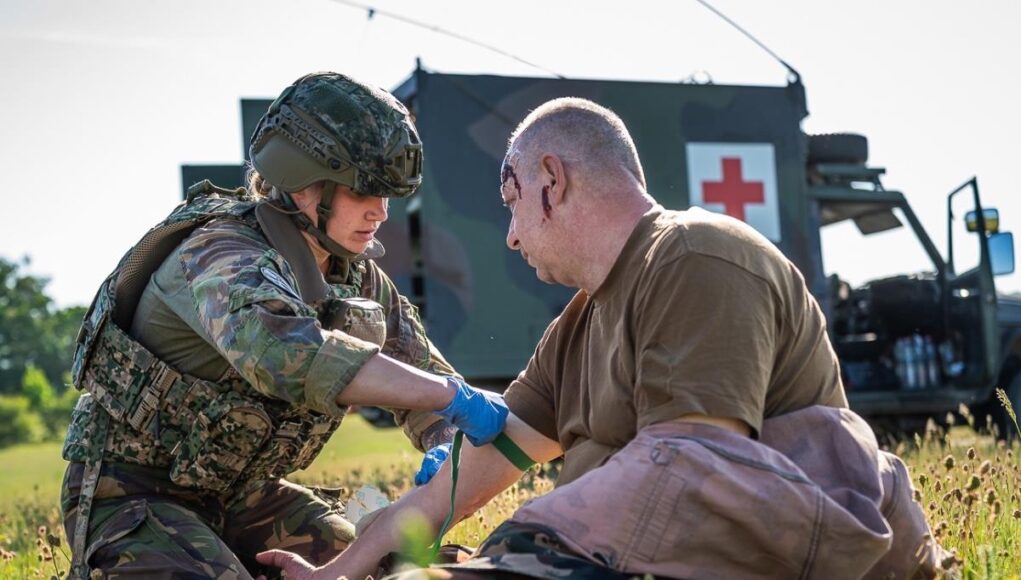Oppus-Vigorous-Warrior-2024-Exercise-Vigorous-Warrior-2024 Military medic in camouflage armor bandages a wounded soldier on a grassy field, with a field medical vehicle (Red Cross) in the background.