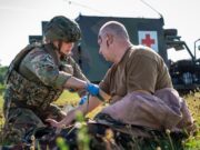NATO holds huge medical drill in Estonia Military medic in camouflage armor bandages a wounded soldier on a grassy field, with a field medical vehicle (Red Cross) in the background.