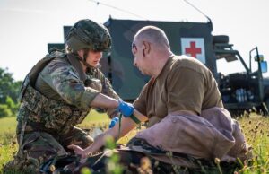 NATO holds huge medical drill in Estonia Military medic in camouflage armor bandages a wounded soldier on a grassy field, with a field medical vehicle (Red Cross) in the background.