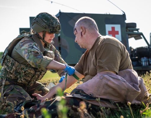 NATO holds huge medical drill in Estonia Military medic in camouflage armor bandages a wounded soldier on a grassy field, with a field medical vehicle (Red Cross) in the background.