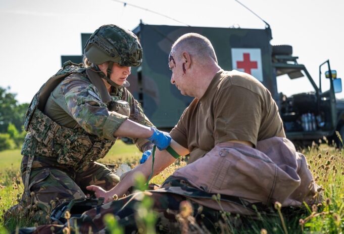 Military medic in camouflage armor bandages a wounded soldier on a grassy field, with a field medical vehicle (Red Cross) in the background.