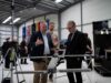 Three men in a workshop inspect a large drone mounted on a stand, with flags along the wall in the background.
