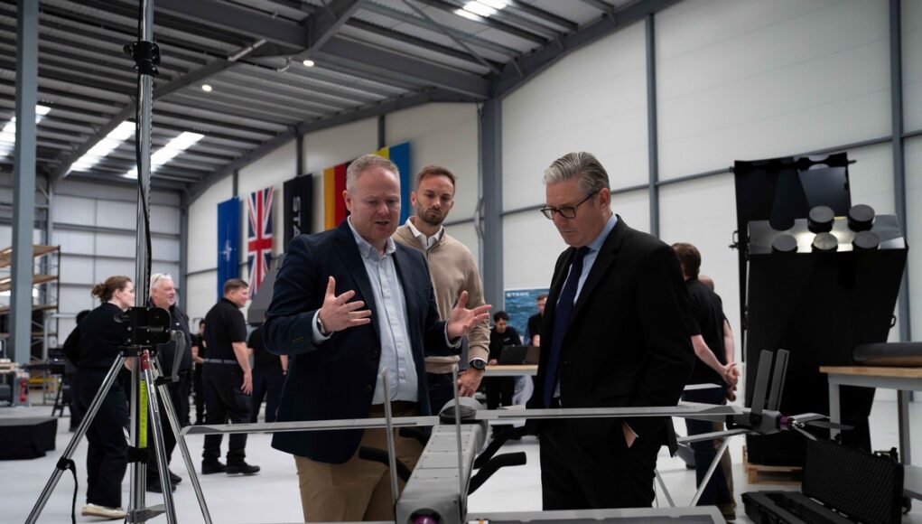 STARK - PM visit Three men in a workshop inspect a large drone mounted on a stand, with flags along the wall in the background.