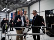 STARK debuts Cascade and Gambit drones with UK partner Three men in a workshop inspect a large drone mounted on a stand, with flags along the wall in the background.