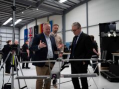 STARK debuts Cascade and Gambit drones with UK partner Three men in a workshop inspect a large drone mounted on a stand, with flags along the wall in the background.