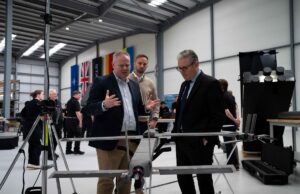 STARK debuts Cascade and Gambit drones with UK partner Three men in a workshop inspect a large drone mounted on a stand, with flags along the wall in the background.