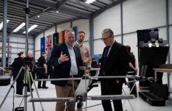 STARK debuts Cascade and Gambit drones with UK partner Three men in a workshop inspect a large drone mounted on a stand, with flags along the wall in the background.