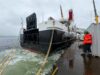 Large vessel docked at a quay with thick mooring lines; a worker in a high-visibility jacket and hard hat stands on the dock nearby.